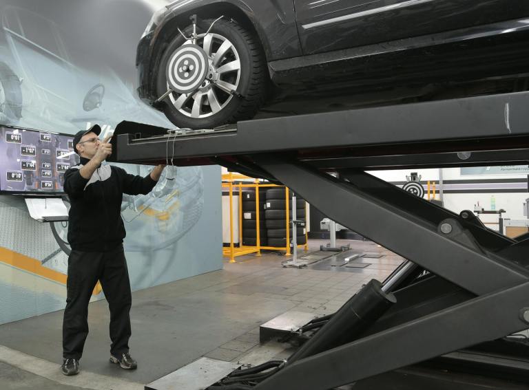A mechanic lifts a car on a hydraulic lift in an automotive workshop, adjusting a wheel while surrounded by tools and equipment.
