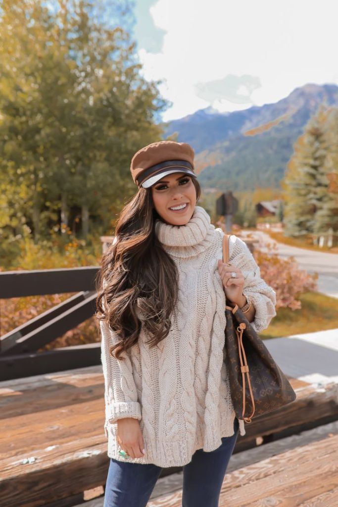 A woman wearing a cozy cream cable-knit sweater, blue jeans, and a brown cap, posing outdoors with autumn foliage and mountains in the background.
