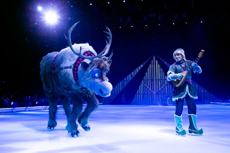 A performer dressed as Kristoff plays a musical instrument alongside a reindeer on an ice rink, with colorful stage lighting in the background.