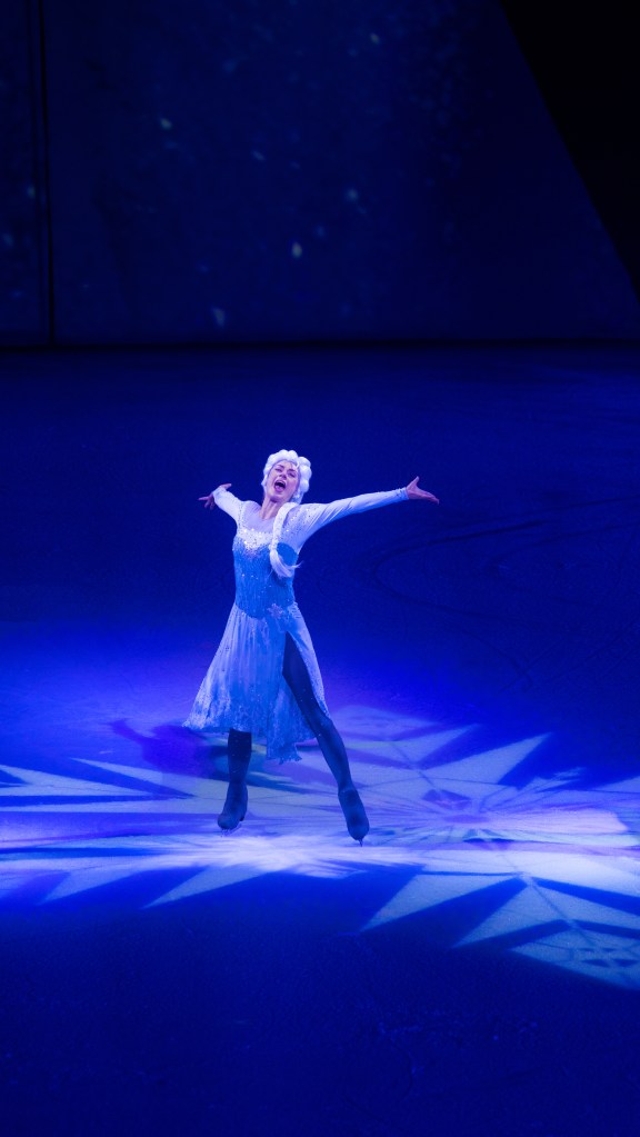 A performer portraying Elsa from Frozen, in a sparkling blue costume, skating on ice under dramatic lighting.