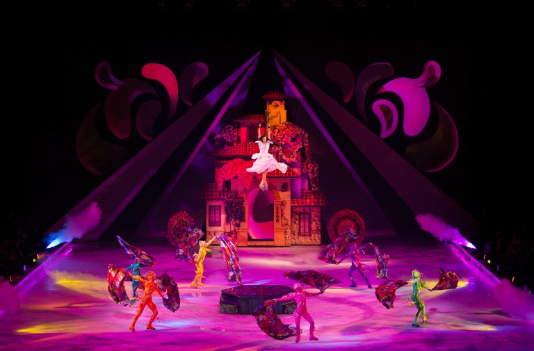 A colorful ice skating performance featuring a dancer in a white costume soaring above the ice surrounded by performers in vibrant costumes, with a visually striking backdrop inspired by the film Encanto.