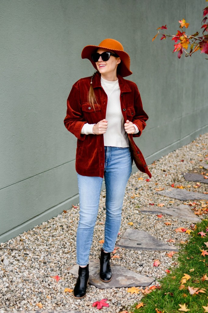 A woman smiling while wearing a warm, rust-colored shacket, paired with a cream sweater and straight-leg jeans. She completes her look with a wide-brimmed orange hat and black ankle boots, standing on a path with autumn leaves around her.