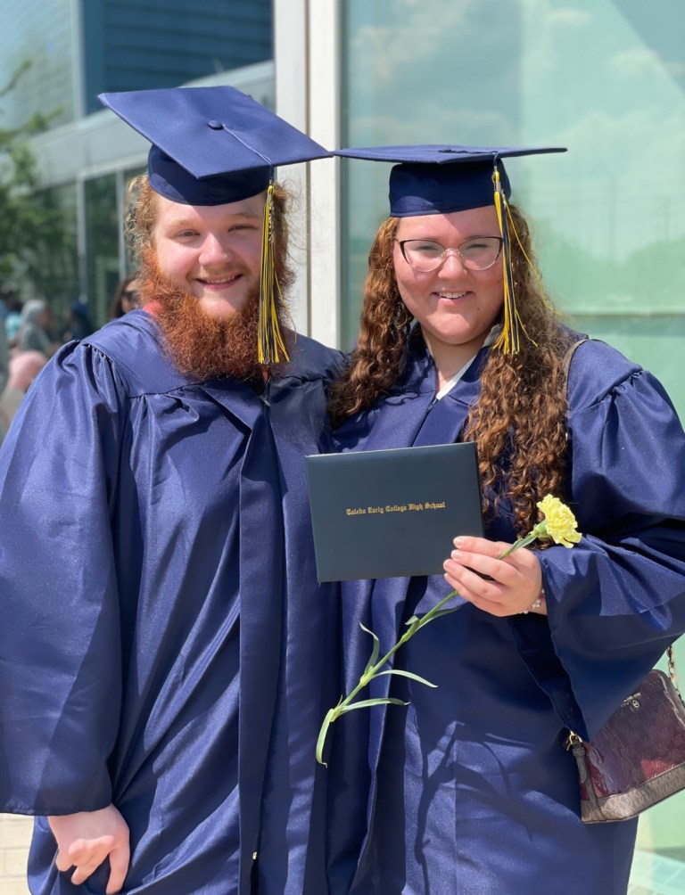 Two high school graduates wearing blue caps and gowns, standing outdoors, one holding a diploma and a yellow flower.
