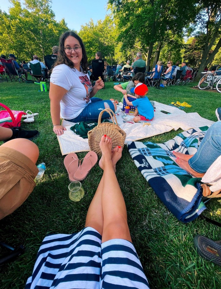 A woman smiling while sitting on a picnic blanket in a park, with her legs crossed and wearing a striped dress. In the background, people are enjoying a gathering, and a child is playing nearby.