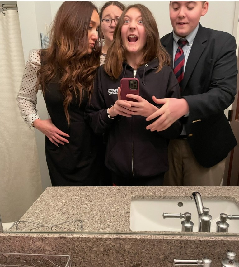 A family of four posing for a selfie in a bathroom mirror, with one child excitedly holding a phone and making a funny face while others strike playful poses.