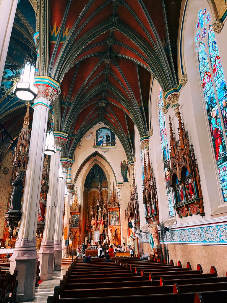 Interior view of a beautifully decorated church, showcasing stained glass windows, intricate architectural details, and wooden pews with people present.