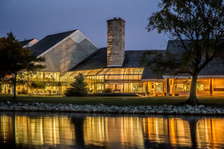 A nighttime view of Maumee Bay Lodge & Conference Center reflecting on the calm waters, showcasing its modern architecture with warm lights glowing from the windows.