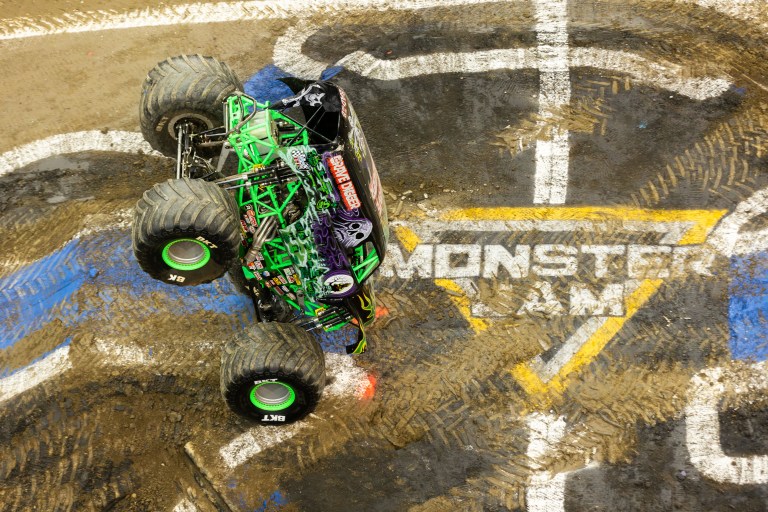 A green and black monster truck performs a stunt on a Monster Jam track, with mud and tire marks visible on the surface.