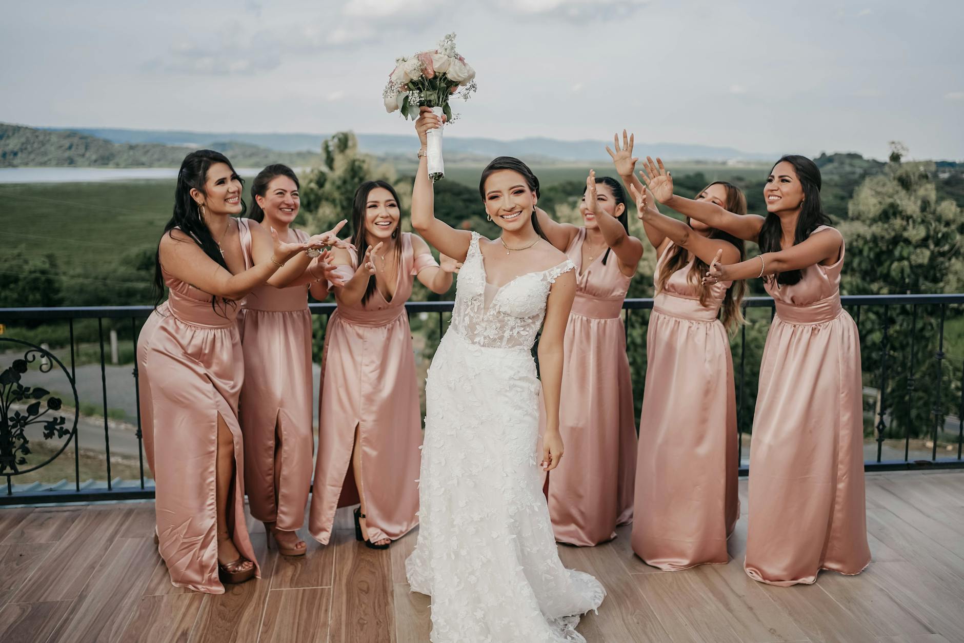 A bride in a white wedding dress joyfully holds a bouquet, surrounded by her bridesmaids in matching pink dresses, celebrating together outdoors.