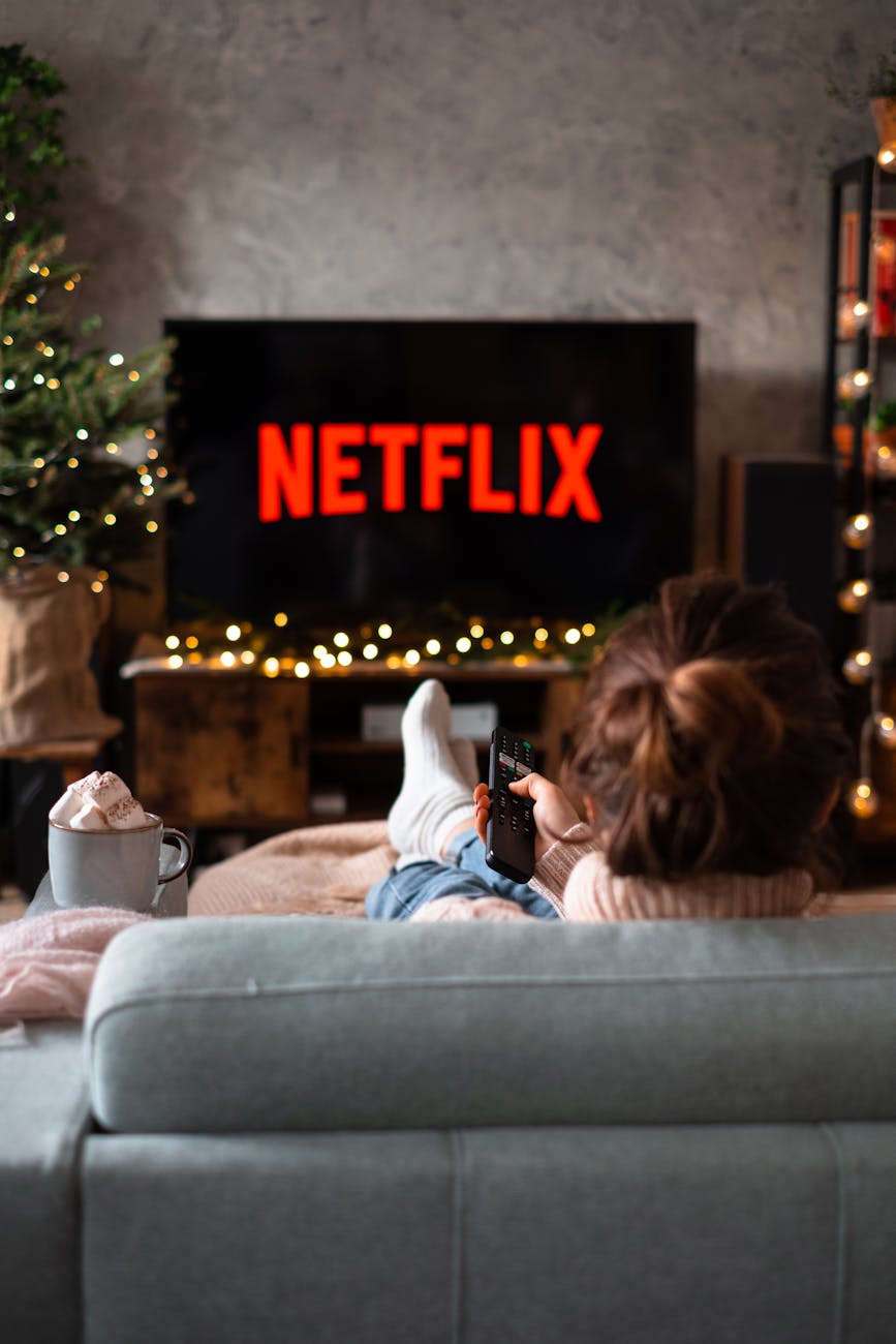A person wrapped in a cozy sweater, relaxing on a sofa with their feet up, holding a remote control while watching Netflix on a large screen. A cup of hot chocolate with marshmallows is placed next to them, and a decorated Christmas tree can be seen in the background.