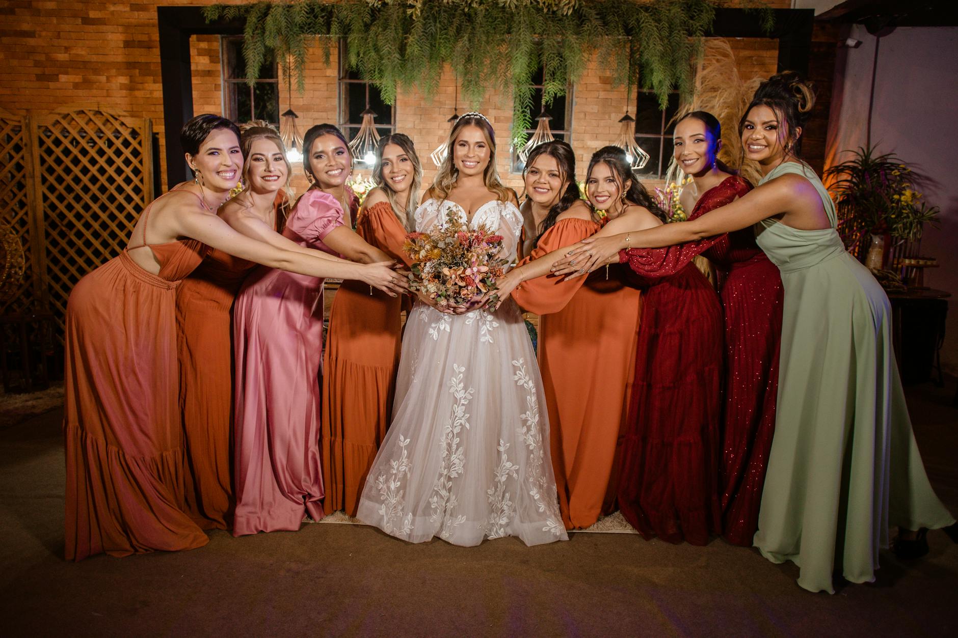 A bride surrounded by her bridesmaids, each wearing dresses in various colors and styles, posing together joyfully in a beautifully decorated venue.