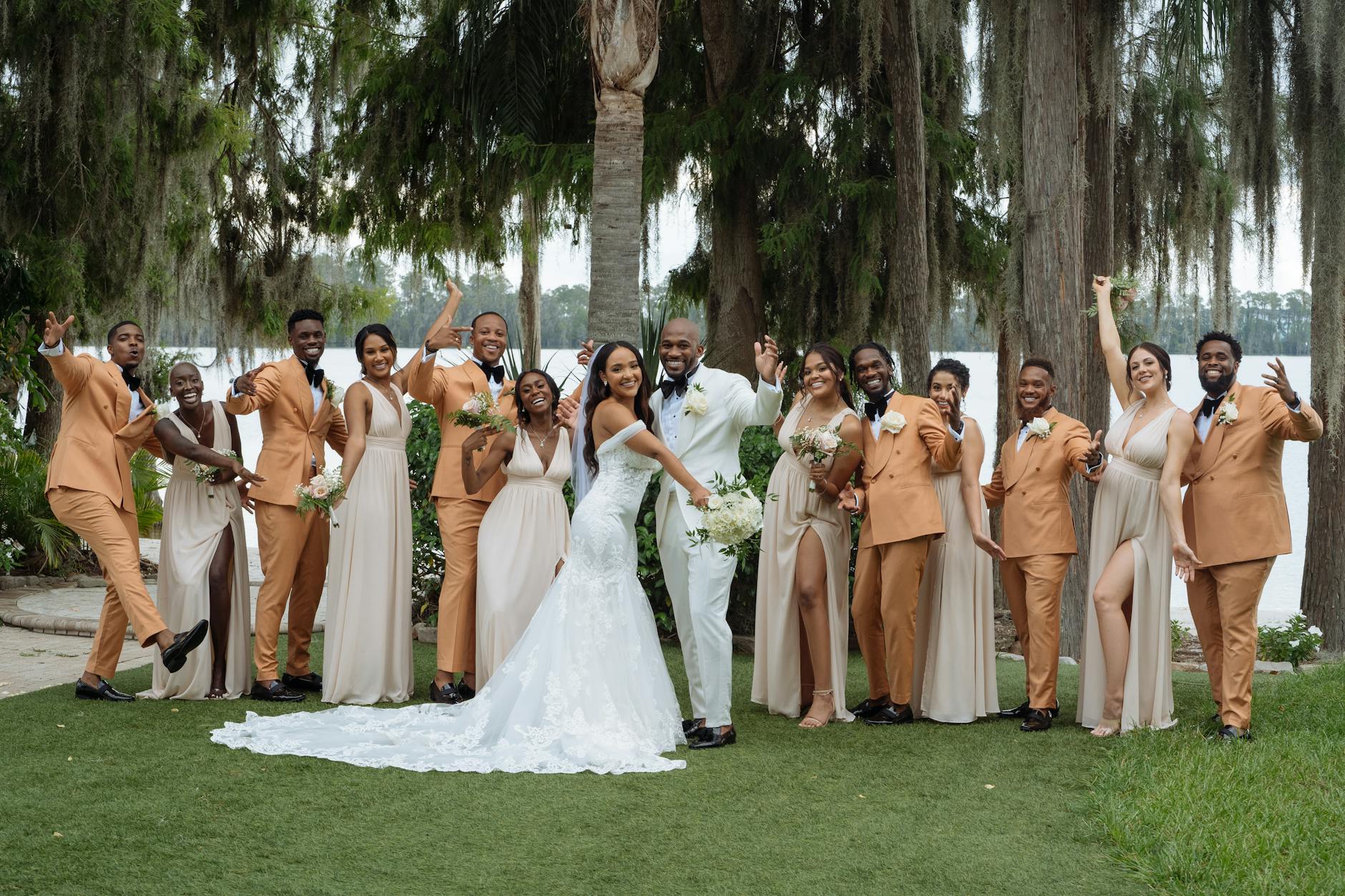 A joyful group of bridesmaids and groomsmen posing together outdoors, featuring a bride in a white wedding gown and her groom in a white suit. The bridesmaids wear light-colored dresses while the groomsmen are dressed in coordinating tan suits.