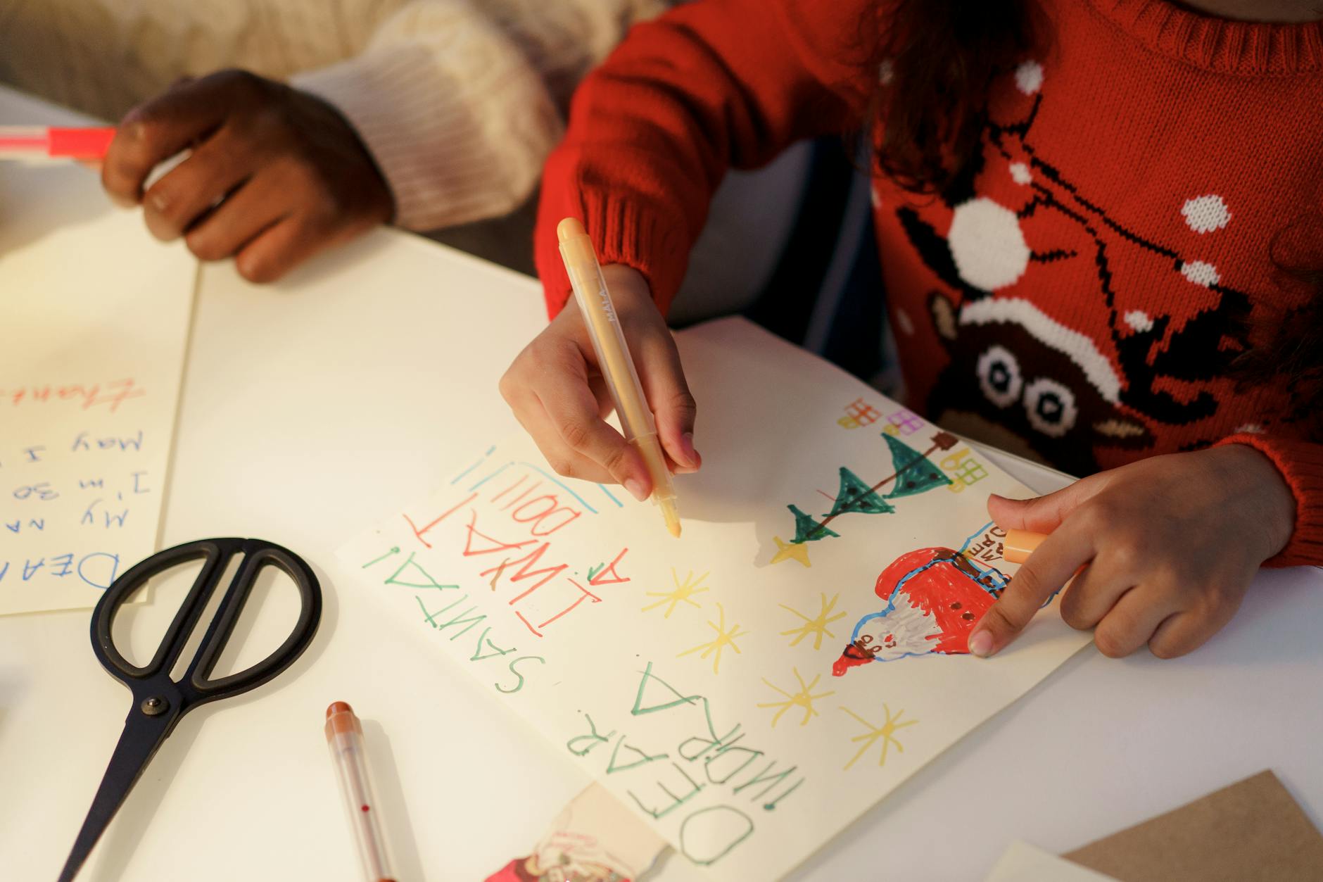 A child in a red holiday sweater is drawing on white paper with colorful markers, working on a festive letter to Santa, surrounded by craft supplies and scissors.