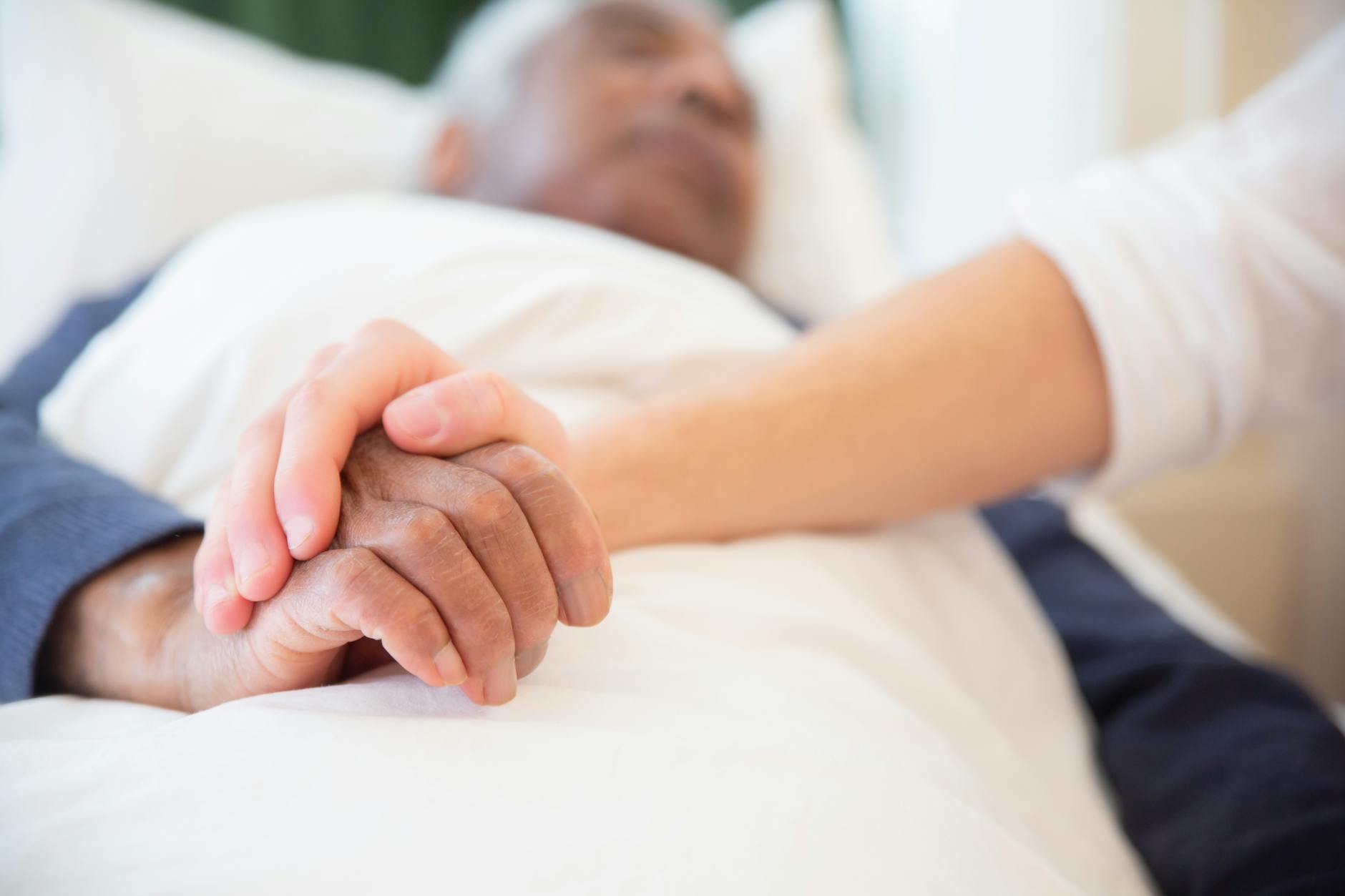 A close-up of two hands intertwined, one with darker skin and the other with lighter skin, symbolizing care and support while a person is resting in bed.