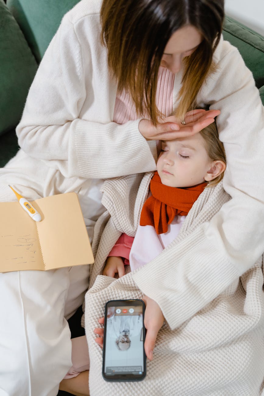 A mother gently checking her child's forehead while the child sleeps, wrapped in a cozy blanket. On a nearby table, a thermometer and a notebook can be seen, while the mother holds a phone displaying a medical consultation.