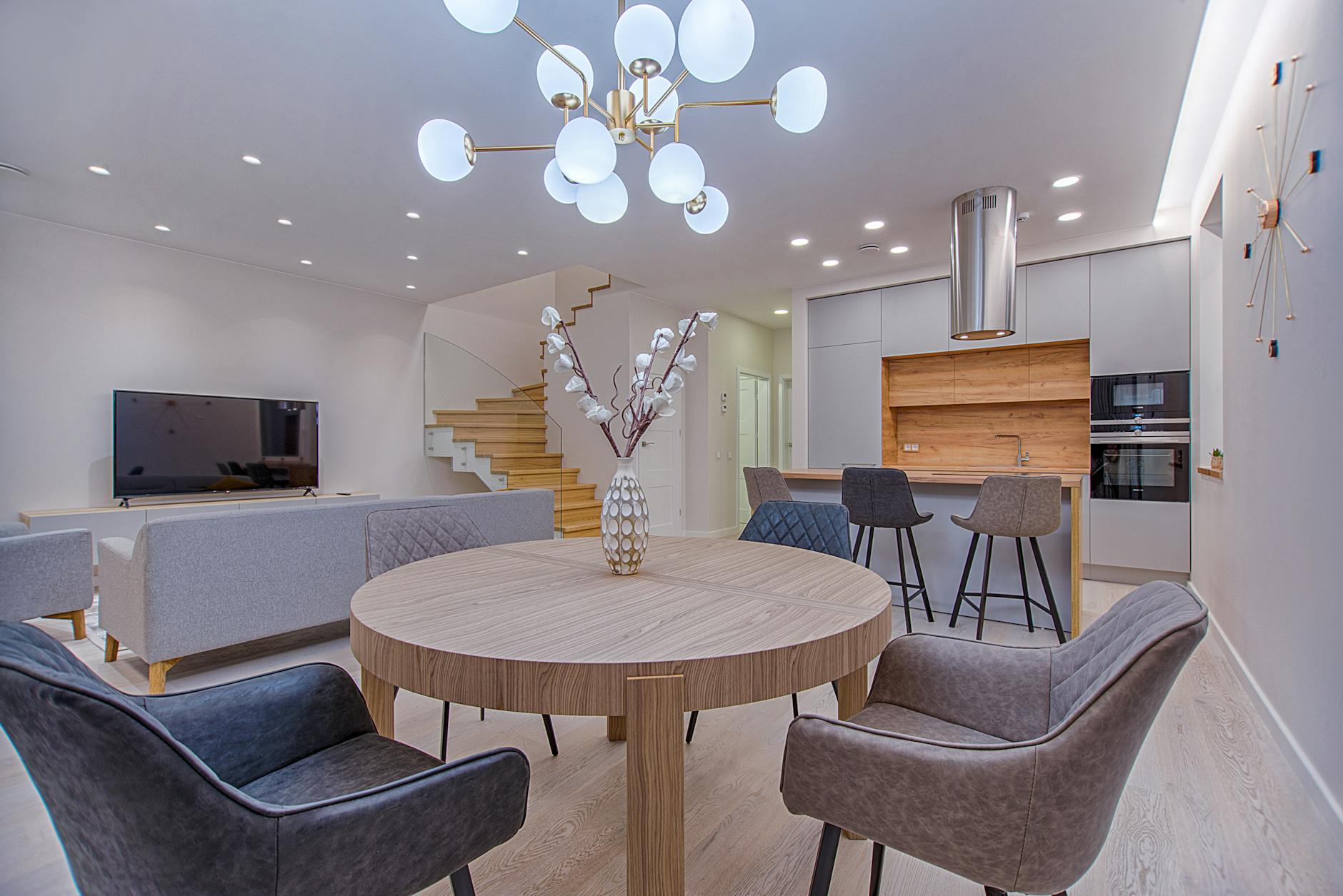 Modern living room and kitchen area featuring a round wooden dining table, chairs, a television, and stylish decor elements.