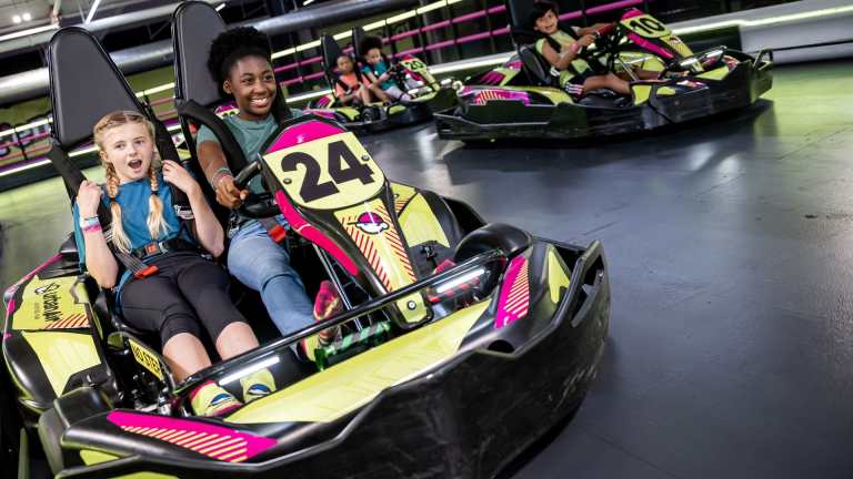 Two children enjoying a go-kart ride, with one girl excitedly screaming and the other smiling, in a brightly colored go-kart track.