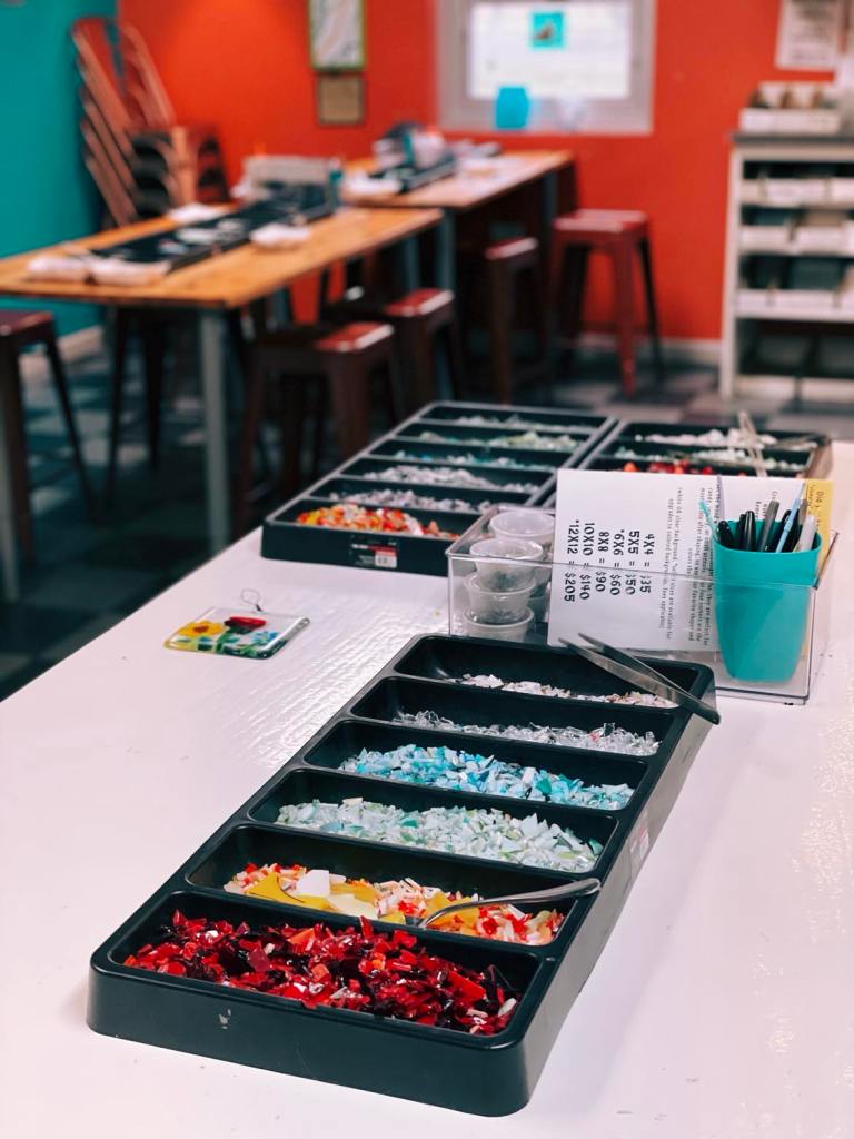 A variety of colored glass pieces arranged in trays on a work table in a studio, with workshop materials and colorful walls in the background.