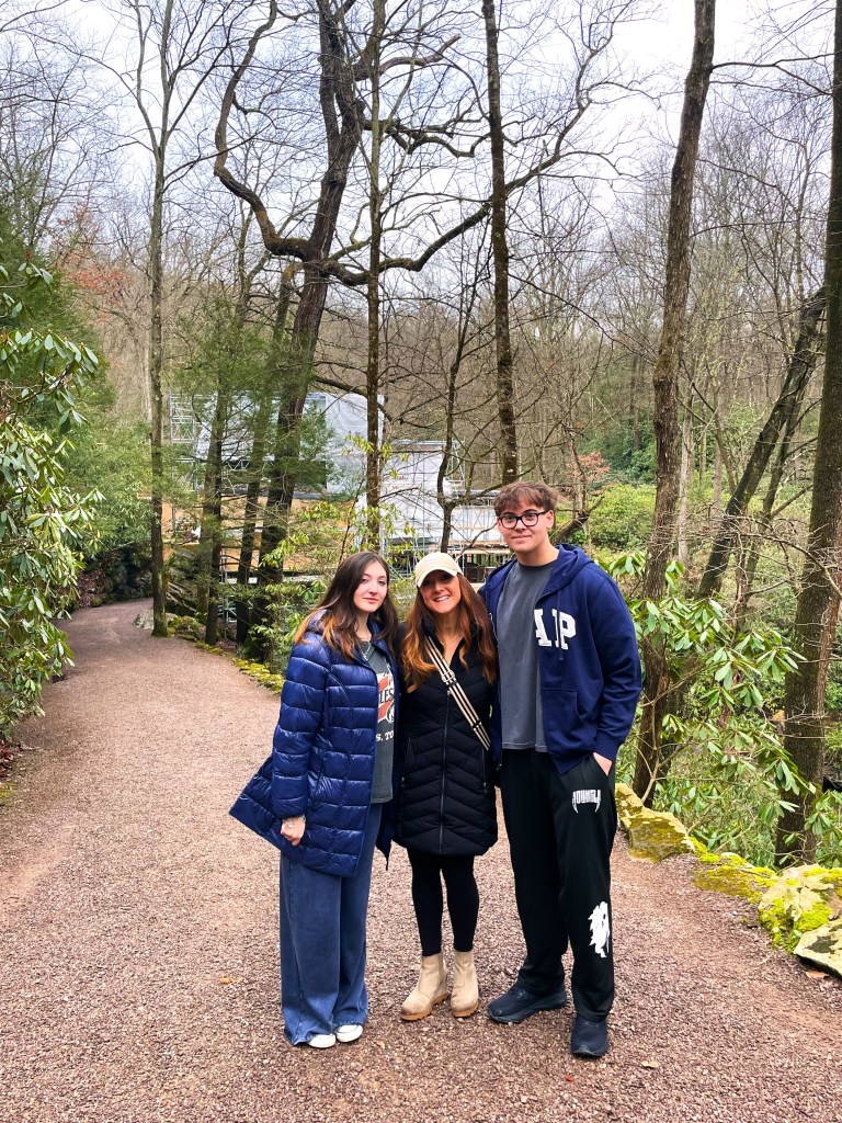 Three individuals standing on a gravel path in a wooded area, surrounded by tall trees and shrubs. They are dressed in winter clothing and smiling at the camera.