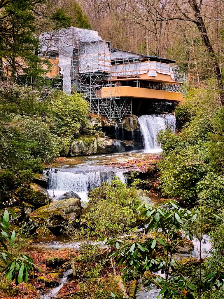 A house under construction surrounded by scaffolding, located next to a flowing waterfall and rocky stream, with trees and greenery in the foreground.