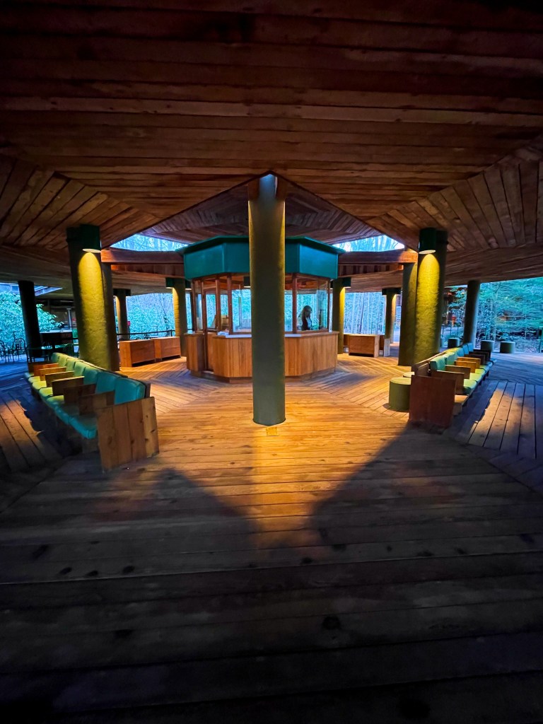 Interior view of a wooden structure featuring a central kiosk and circular seating, illuminated with warm lighting.