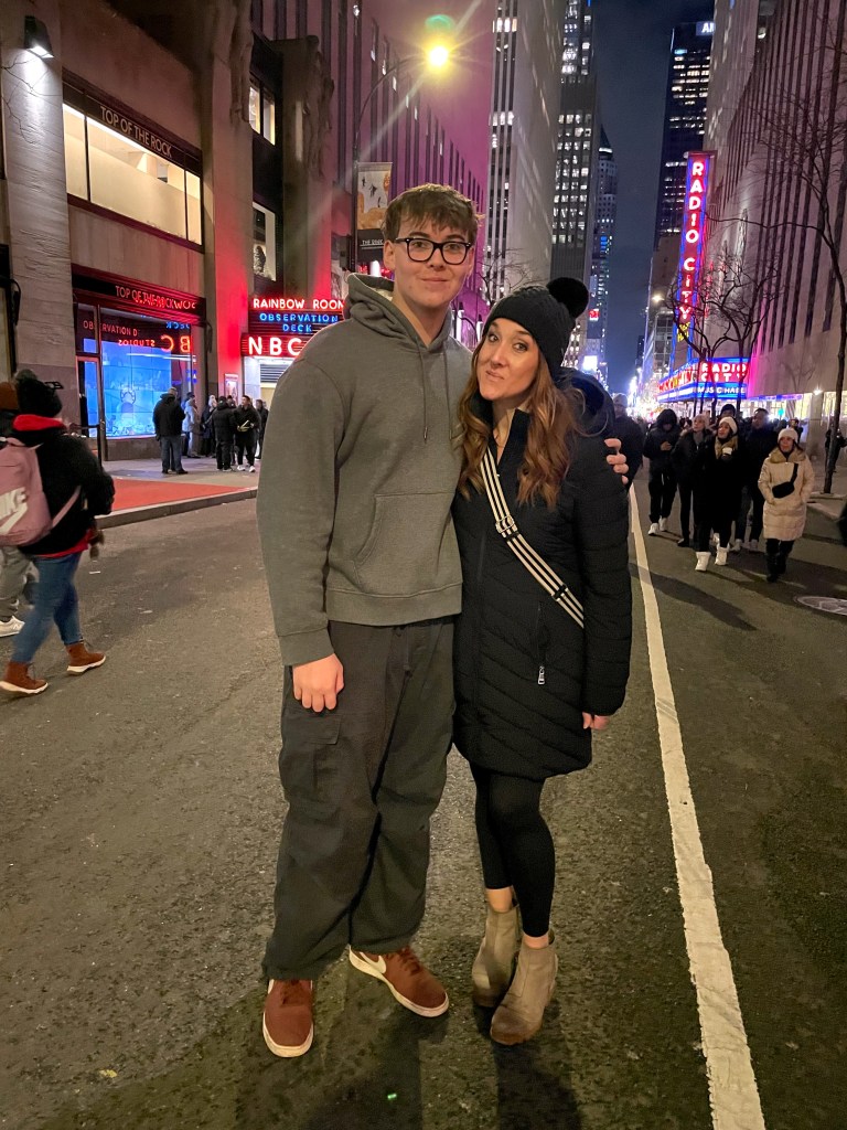 A young couple poses for a photo on a bustling street at night, surrounded by city lights and buildings. The male is wearing a gray hoodie and casual pants, while the female is dressed in a dark coat and hat, smiling together.
