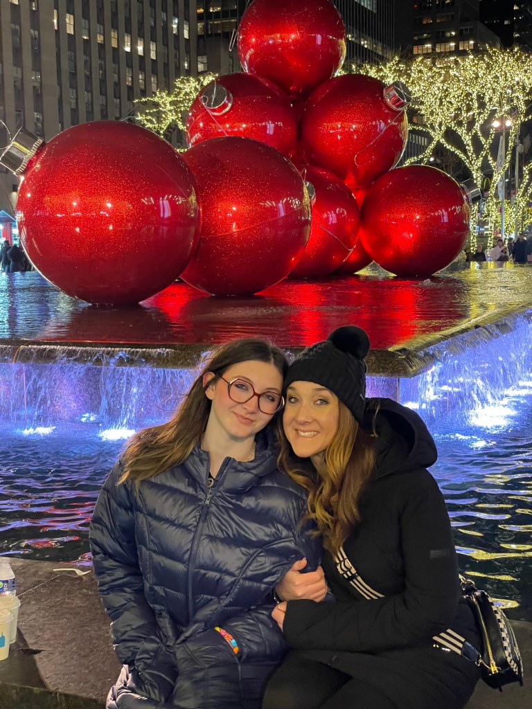 A joyful moment between two women sitting by a fountain adorned with large red Christmas ornaments, illuminated by colorful lights in an urban setting at night.