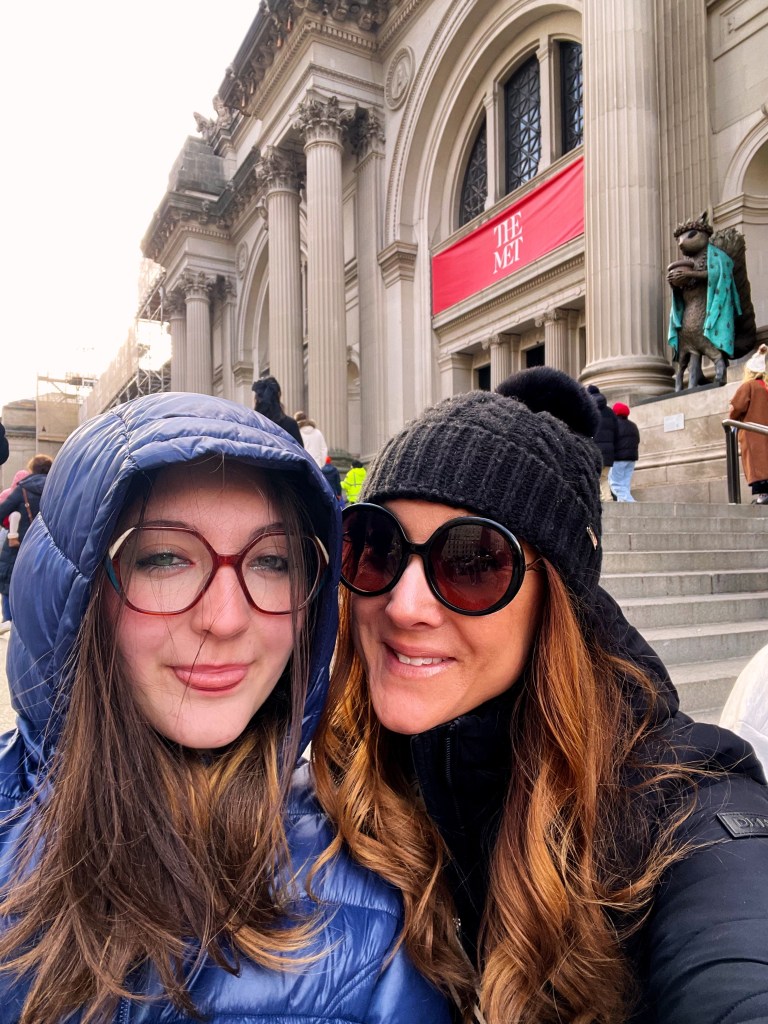 Two women smiling for a selfie in front of The Metropolitan Museum of Art, wearing winter coats and accessories, with the museum's grand facade visible in the background.