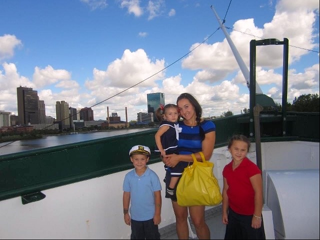A woman holds a child while standing on a boat deck with two other children nearby, overlooking a city skyline and river under a blue sky with clouds.