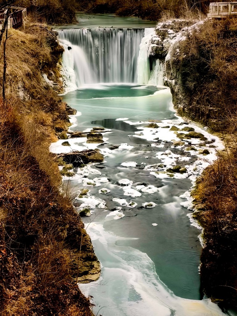 A scenic view of a waterfall cascading into a turquoise river surrounded by rocky formations and sparse vegetation, with patches of ice along the water's edge.