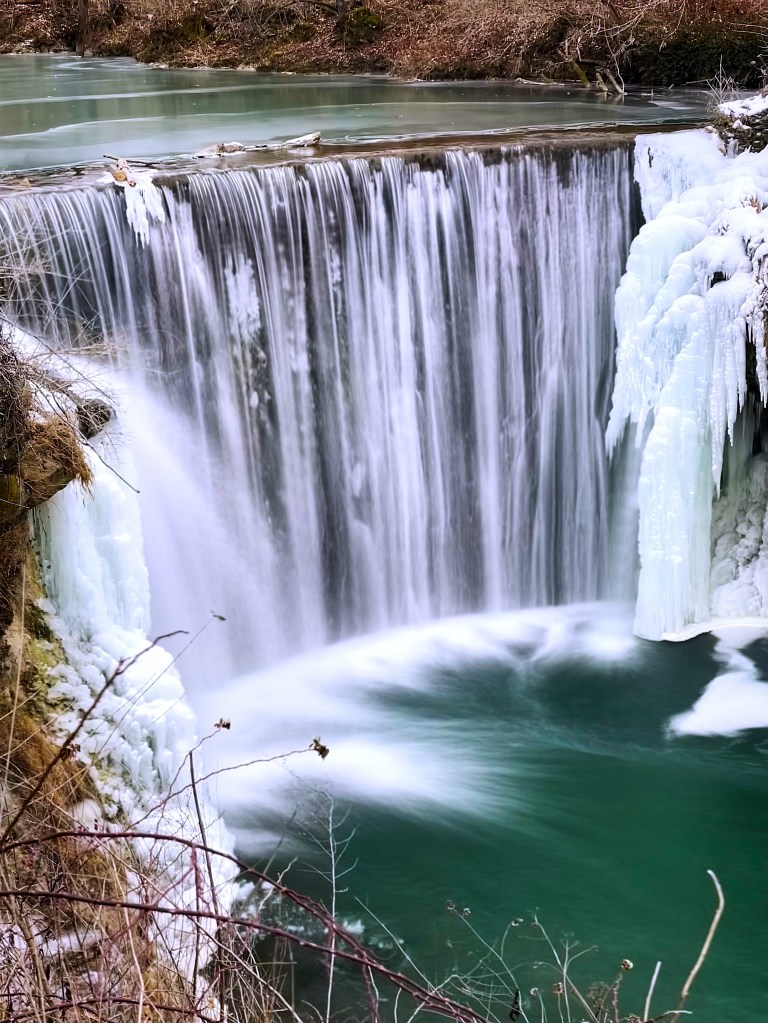 A frozen waterfall cascading into a turquoise pool, surrounded by icy formations and barren branches.