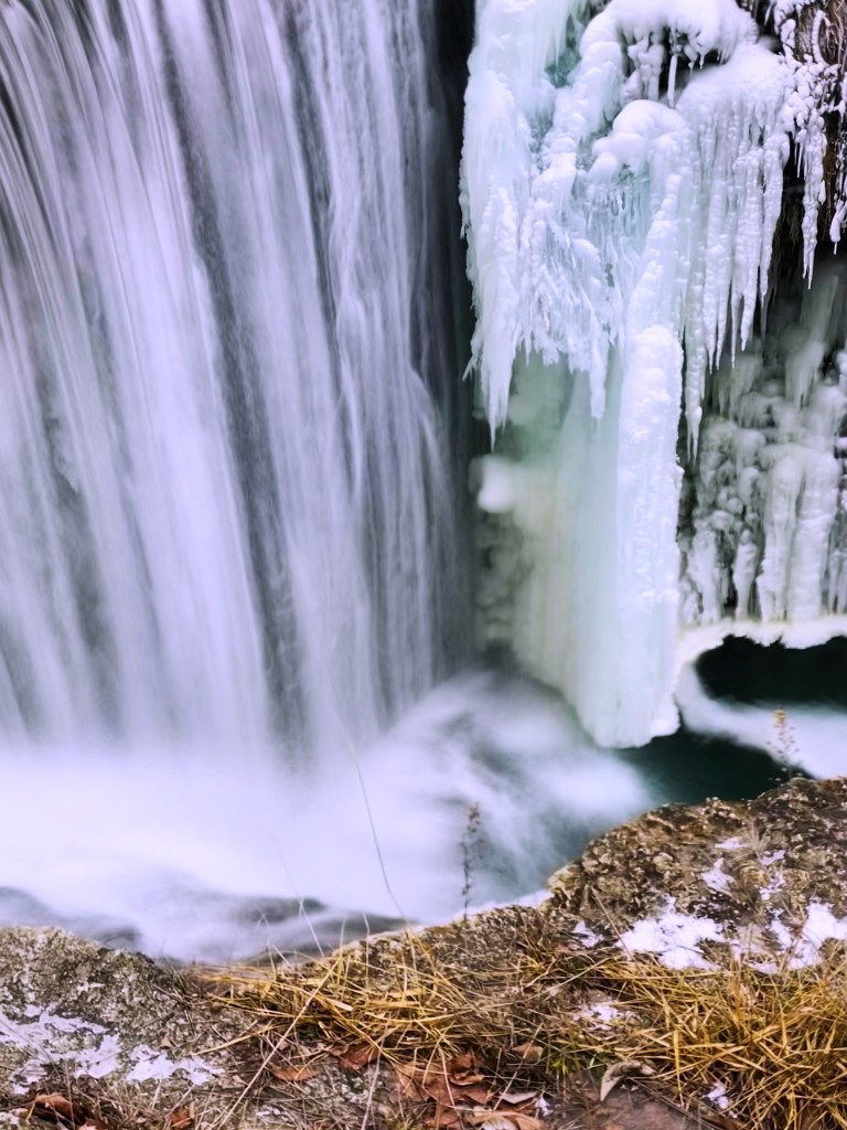 A close-up view of a frozen waterfall with blue ice formations and flowing water at the base, surrounded by a rocky terrain and sparse grass.
