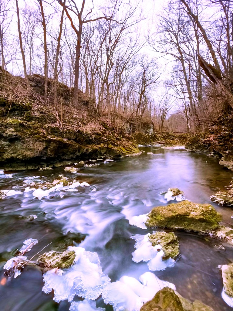 A tranquil stream flowing through a rocky landscape, surrounded by bare trees in early spring. Ice patches are visible on the water's surface, creating a serene natural scene.
