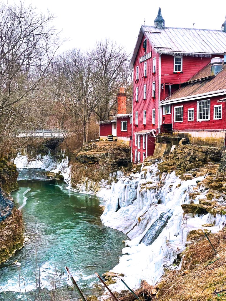 A red historic building beside a frozen waterfall, with icy rocks and a green river, surrounded by bare trees.