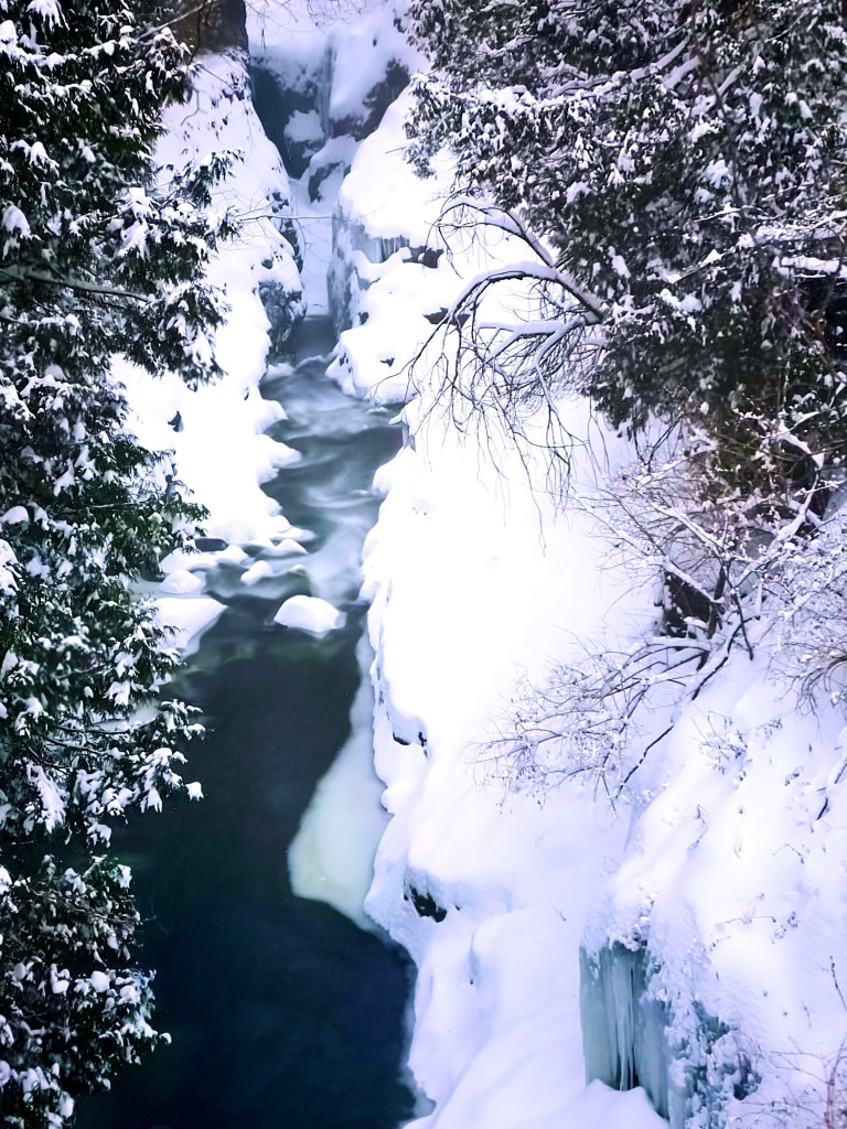 A snow-covered riverbank with steep, icy cliffs on either side, flowing dark water partially obscured by patches of snow and ice.