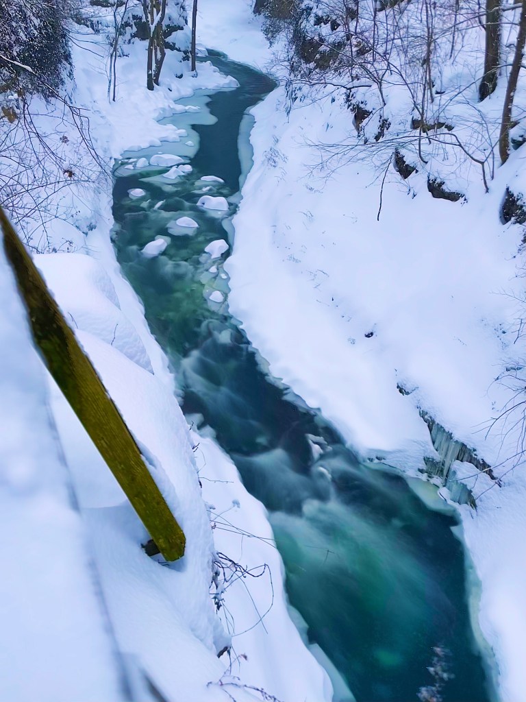 A serene winter landscape featuring a narrow, flowing river surrounded by snow-covered banks and trees. Ice and snow are visible along the edges of the river.