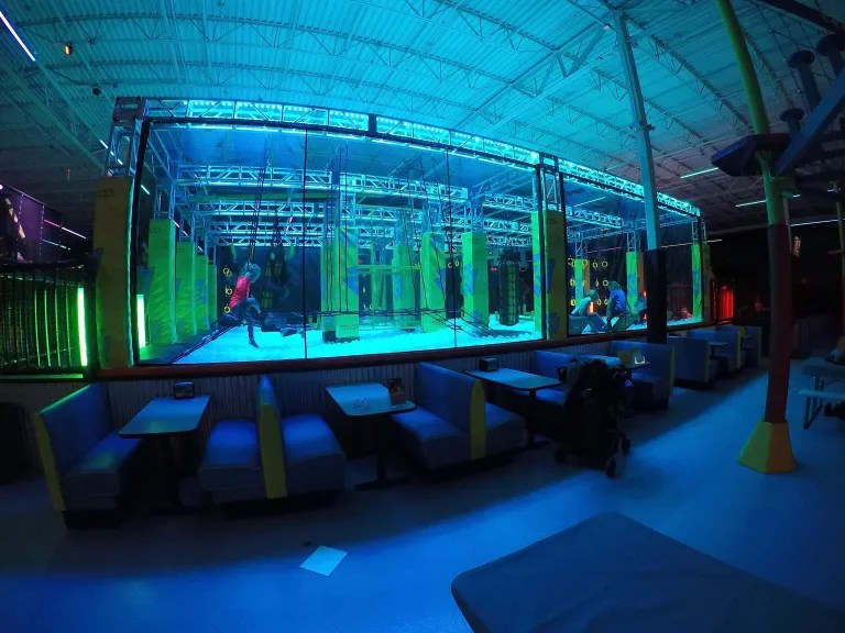 Indoor adventure park featuring a climbing area with colorful walls and obstacles, illuminated with blue and green lights. Seating area with blue booths in the foreground.