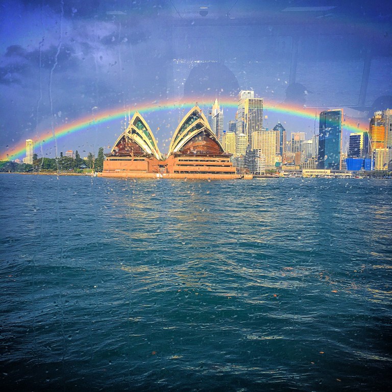 Sydney Opera House under a rainbow with the city skyline in the background, viewed from the water.
