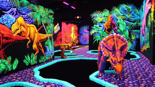 A vibrant blacklight indoor miniature golf course featuring colorful dinosaur murals and three-dimensional dinosaur sculptures among lush tropical vegetation.