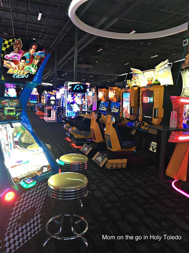 An indoor arcade filled with various video game machines, racing simulators, and colorful lights. Stools are situated in front of some machines, creating a vibrant gaming atmosphere.