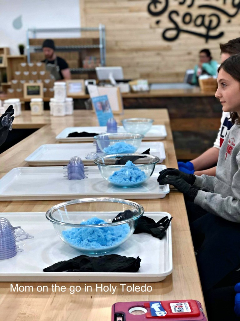 A workshop setup for a soap-making class featuring bowls of blue soap mixture, black gloves, and participants preparing their materials at a workspace.