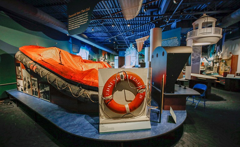 A close-up view of an orange lifeboat and a red life ring displayed in a maritime museum exhibit, featuring various nautical artifacts in the background.