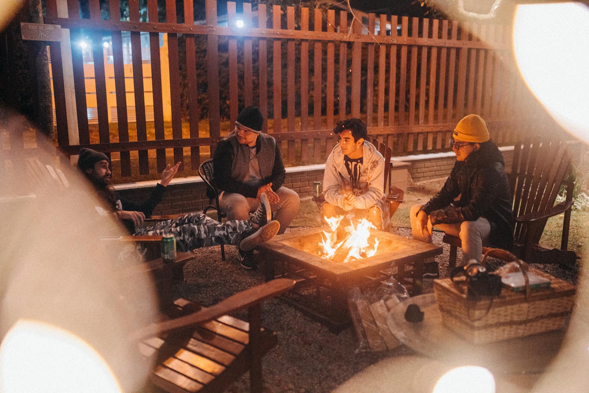 Group of four people sitting around a fire pit at night, engaged in conversation. The ambiance is cozy with wooden seating and a wooden fence in the background.