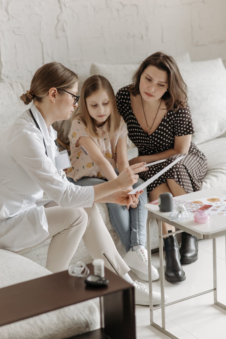 A healthcare professional explains something to a young girl and her mother while sitting on a couch, with medical materials and a tablet on a table in front of them.