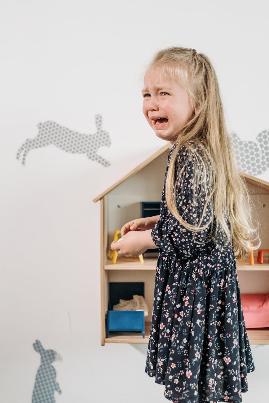 A young girl with long blonde hair is crying while holding a small toy in a playroom decorated with rabbit silhouettes.