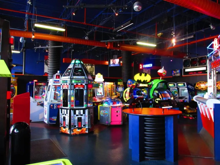 Interior view of an arcade featuring various colorful gaming machines, including racing games and skill challenges, under vibrant lighting.