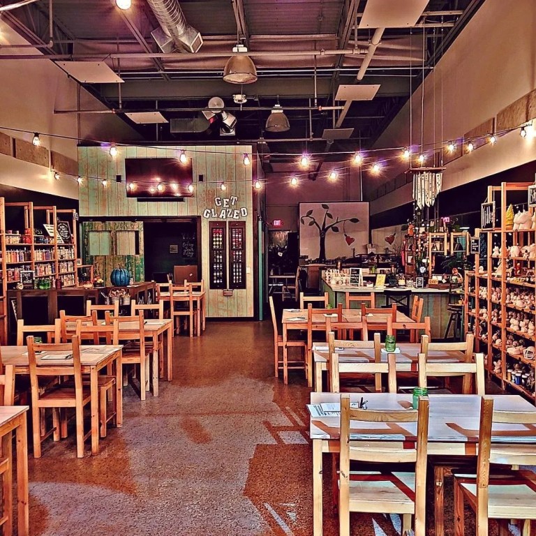 Interior view of a pottery painting studio with wooden tables and chairs, shelves displaying pottery items, and a 'Get Glazed' sign on the wall.