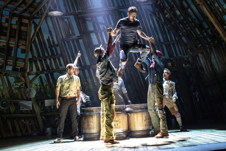 A theatrical performance showing several actors on stage. One actor is being lifted in the air by two others, while additional actors are positioned around them in a barn-like setting with wooden beams and barrels.