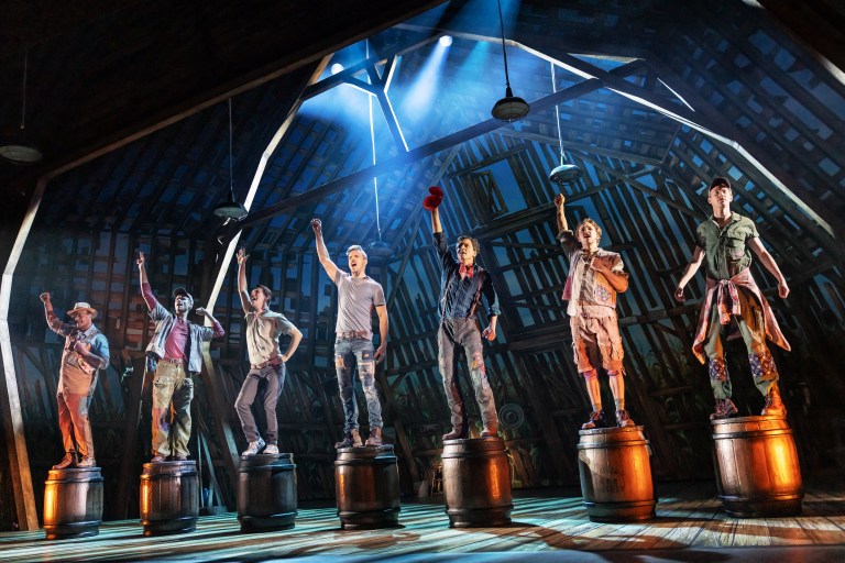 Seven male performers standing on barrels in a barn setting, raising their fists in a show of unity and strength, with dramatic lighting illuminating the scene.
