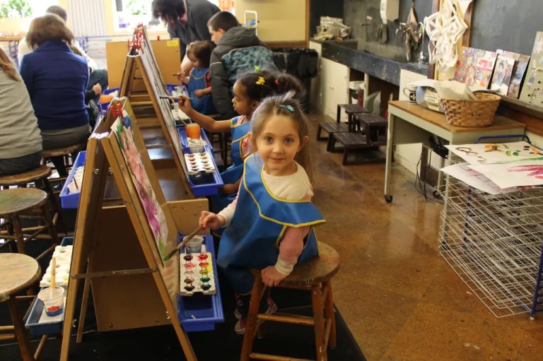 A young girl sitting on a stool in an art class, wearing a blue apron and smiling at the camera, with paint palettes and easels in the background.